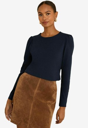 Navy Blaue Langarm-Bluse mit gepufften Schultern, kombiniert mit einem braunen Wildlederrock mit Mittelnaht und weicher Textur.