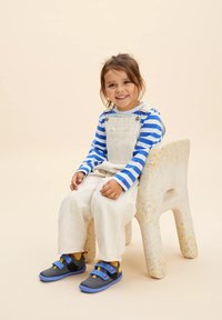 Camisa de manga larga a rayas azul y blanca, tirantes beige y zapatos grises con correas azules. Una silla de color claro en un fondo neutro.
