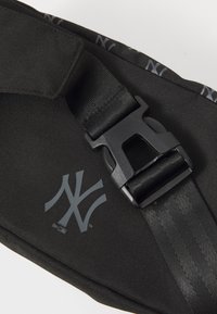 Sac en tissu noir avec une texture lisse, arborant un logo gris, une sangle épaisse et des détails en plastique noir.