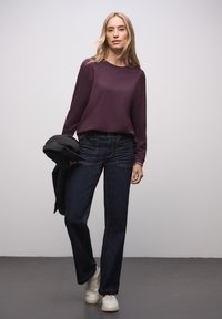 Donkerblauwe jeans met rechte pijpen gecombineerd met een diep paarse top met lange mouwen. Witte sneakers en een zwart jasje worden nonchalant over de arm gedragen.