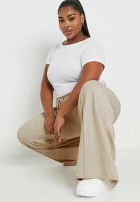 Valkoinen istuva t-paita ja beige leveälahkeinen housu, jossa on nauhakiristyksellä varustettu vyötärö. Malli poseeraa polvillaan, jalassaan valkoiset tennarit. Sujuva kankaan rakenne.