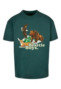 Zöld pamut póló, amelyen egy medve, szarvas és sas grafikája látható, a "Beastie Boys" felirattal, mely világos sárga betűkkel van kiemelve.