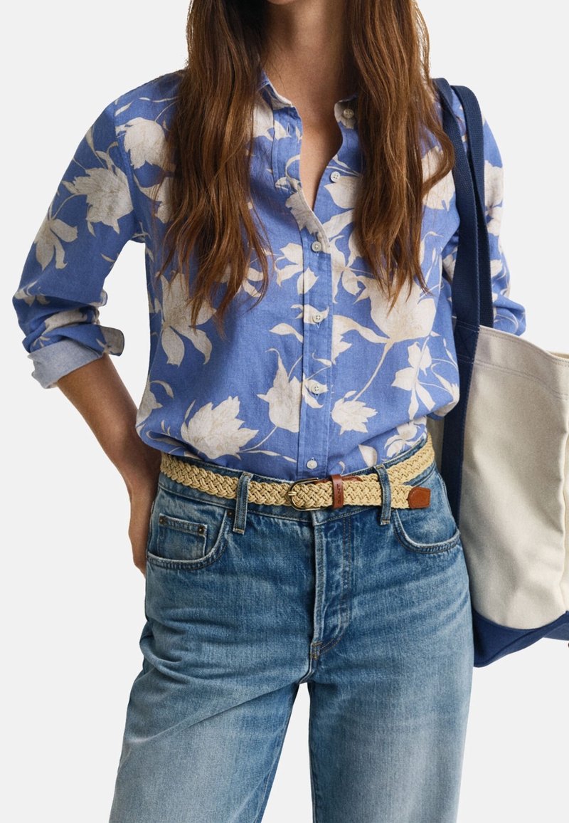 Femme portant une chemise bleue à fleurs boutonnée avec les manches retroussées, une ceinture tissée claire, un jean bleu et un sac cabas beige et bleu marine porté à l'épaule.