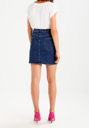 Kvinde iført en hvid kortærmet top, mørkeblå denim nederdel og lyserøde høje hæle, stående med ryggen mod kameraet på en hvid baggrund.