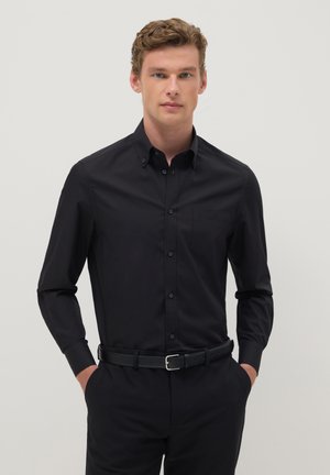 Junger Mann mit lockigem Haar, trägt ein schwarzes Hemd mit Knöpfen und schwarze Hose, die Hände in den Taschen, vor einfarbigem Hintergrund.