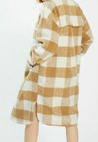 Randig översized kappa i beige och krämfärgad, tillverkad av mjukt tyg. Har sidslitsar och en avslappnad passform med en krage.