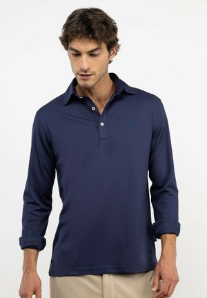 Hombre con polo azul marino de manga larga con dos botones, pantalones beige, mangas remangadas, mirando hacia abajo a su izquierda.