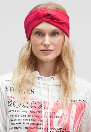 Red fabric headband with a twisted design, made of soft material. Worn by a model with long blonde hair, also wearing a white sweater.