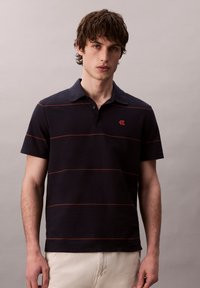 Námořnická polokošile s krátkými rukávy, červenými vodorovnými pruhy a malým vyšitým logem na hrudi. Bavlněná tkanina a klasický design límce.