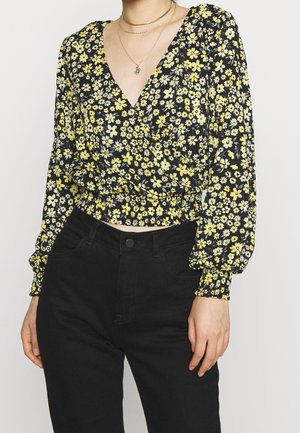Femme portant un crop top à manches longues noir à motifs floraux jaunes et blancs, avec un pantalon noir taille haute, montrant le décolleté et les bijoux.