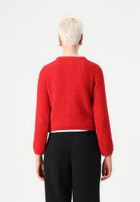 Pull en maille texturée rouge avec un col rond et des manches longues, coupe courte, associé à un pantalon noir taille haute.