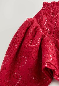 Czerwona bluzka z koronką, charakteryzująca się bufiastymi rękawami i kwiatowymi wzorami. Materiał zdobiony jest cekinami, tworząc teksturowany, dekoracyjny wygląd.