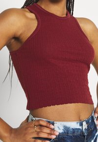 Burgundský žebrovaný crop top s vysokým výstřihem a bezrukávovým designem, který má přiléhavý střih a surový, nepravidelný lem.