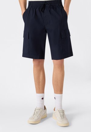 Mężczyzna ubrany w czarne krótkie spodnie typu cargo z bocznymi kieszeniami, białe skarpetki i kremowe sneakersy, stoi z rękami w kieszeniach.