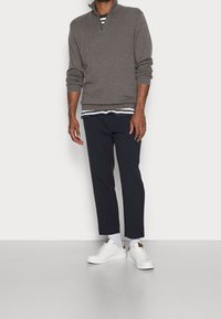Maglione grigio con collo a zip sopra una camicia navy a righe, abbinato a pantaloni navy affusolati e sneakers bianche con dettagli marroni.