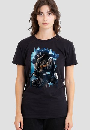 Frau trägt ein schwarzes T-Shirt mit einer Grafik von Batman in Aktionspose vor blauem Hintergrund.