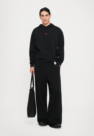 Sweat à capuche noir avec logo rouge à l'avant, cordon de serrage et poignets côtelés. Associé à un pantalon noir à jambes larges et un sac fourre-tout noir. Tissu lisse.