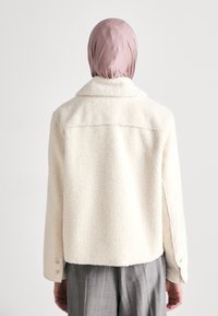 Cremefarbene Fleecejacke mit weicher Textur, Kragen und Knopfdetails an den Ärmeln, von hinten betrachtet.