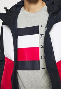 Chaqueta tipo plumón en navy, rojo y blanco con un cuello alto; llevada sobre una camiseta gris claro con un diseño de logo geométrico en negro y rojo.