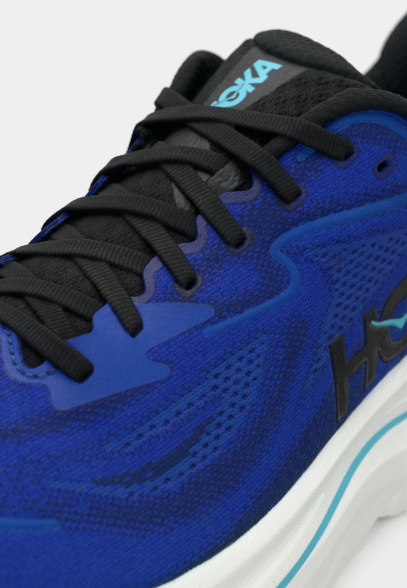 Närbild av en blå och svart löparsko med svarta snören, ventilerande mesh-övre och vit sula med blå randdetaljer.