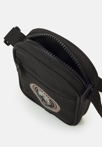 Czarna torebka na ramię z płótna z zamkiem błyskawicznym, z przednim naszywką i regulowanym paskiem. Kompaktowy kwadratowy kształt o teksturowanej powierzchni.