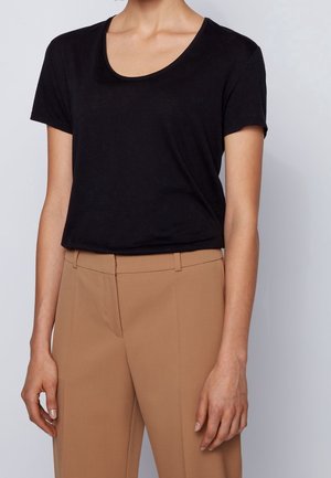 Vrouw die een zwart kortemouwen top met een ronde hals draagt, ingestopt in een camelkleurige, op maat gemaakte broek met hoge taille, tegen een effen achtergrond.