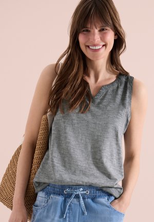 Femme souriante aux cheveux bruns, portant un haut gris sans manches et un pantalon bleu avec cordon, tenant un sac tissé sur son épaule.