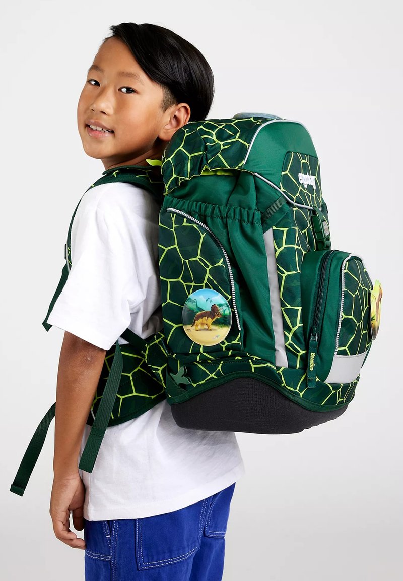 Junge trägt weißes Hemd und blaue Hose, hat einen großen grünen Rucksack mit gelbem Netzmuster und Löwen-Abzeichen an der Seitentasche.