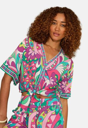 Frau mit lockigem Haar, die ein lebhaftes, farbenfrohes, gemustertes Kurzarmoberteil trägt, das an der Taille gebunden ist, und passende Hosen vor einfarbigem Hintergrund.