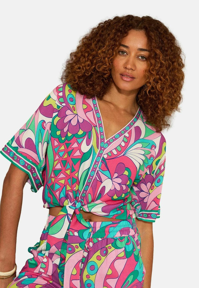 Nainen, jolla on kiharat hiukset, pukeutunut kirkkaanväriseen ja kuvioituun lyhythihaiseen yläosaan, joka on sidottu vyötärölle, ja siihen sopiviin housuihin yksiväristä taustaa vasten.