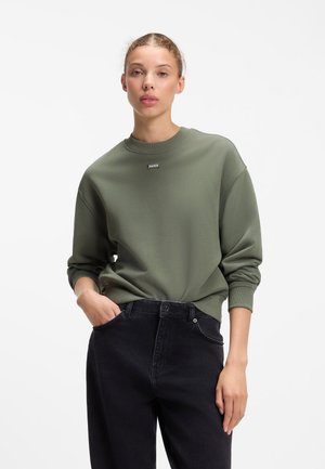 Ung kvinde med lys hud og flettet hår har en grøn Hugo-sweatshirt og sorte jeans på foran en ensfarvet hvid baggrund.