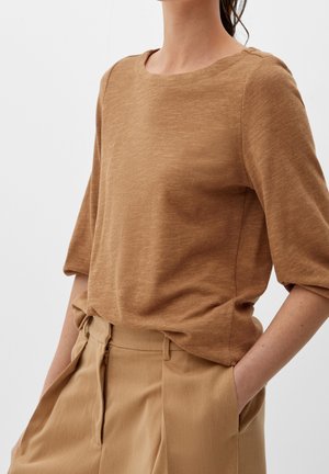 Vrouw die een lichtbruine driekwart mouw blouse draagt, ingestopt in beige broek met hoge taille, handen in de zakken, tegen een effen achtergrond.
