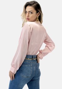 Blusa di raso rosa con maniche lunghe e polsini arricciati, abbinata a jeans a vita alta blu con dettagli di cintura a catena.