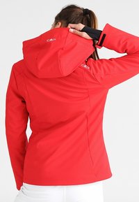 Rød udendørsjakke med tekstureret mønster, fast hætte og tommelhuller, designet til komfort og bevægelsesfrihed. Har logo-accenter.