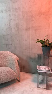 Geschwungener beiger, strukturierter Sessel neben einem Beistelltisch aus Metalldraht mit einer Topfpflanze und gestapelten Zeitschriften, vor einer Betonwand.