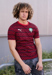 Rød kortærmet trøje med sorte geometriske mønstre, der har et grønt emblem og et hvidt Puma-logo på brystet. Tekstureret stof.