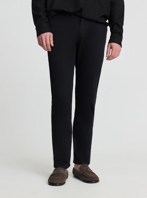 Mężczyzna ubrany w wąskie czarne spodnie, czarną koszulę z długim rękawem i brązowe zamszowe mokasyny, stojący na jednolitym jasnoszarym tle.