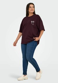 Overdimensjonert burgunder t-skjorte med grafikk av en hvit sløyfe, kombinert med blå smale jeans og kremfargede slip-on sko. Glatt stofftekstur.