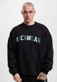 Musta huppari, jonka rinnassa on näyttävä "ROCAWEAR" -teksti valkoisena ja teal-värisenä. Hupparissa on pyöreä kaula-aukko ja ribbikuvioidut hihansuut, ja se on valmistettu pehmeästä kankaasta.