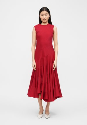 Woman with long black hair wearing a sleeveless red midi dress with an asymmetrical ruffled hem and white pointed-toe shoes.