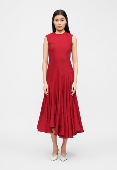 Femme aux longs cheveux noirs portant une robe midi rouge sans manches avec un ourlet asymétrique à volants et des chaussures blanches à bout pointu.