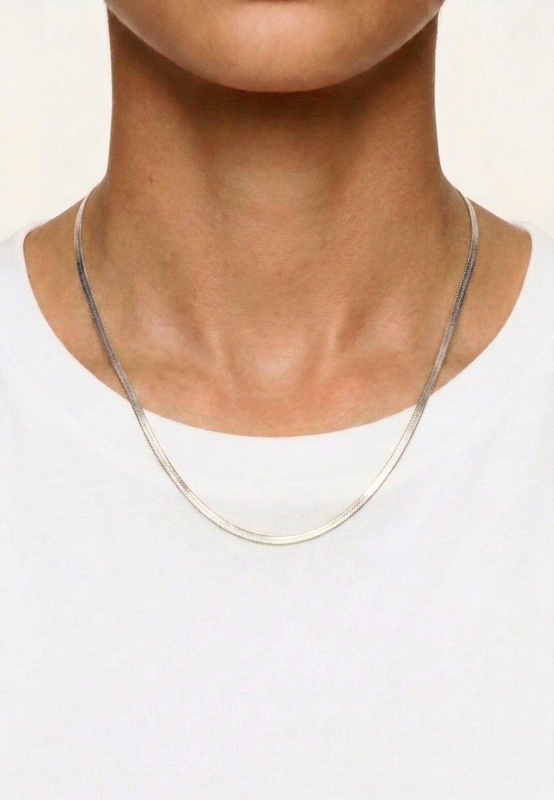 Gros plan d'une personne portant une fine chaîne en argent sur un haut blanc uni, mettant en valeur la zone du cou et de la clavicule.