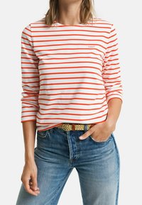 Long-sleeve striped shirt in white and orange, featuring a crew neckline and a small logo. Paired with blue jeans and a braided belt.