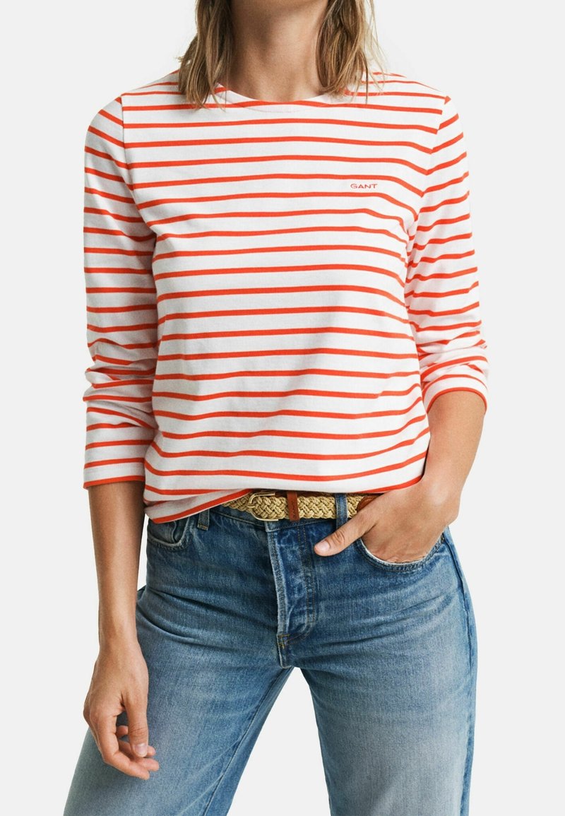 Long-sleeve striped shirt in white and orange, featuring a crew neckline and a small logo. Paired with blue jeans and a braided belt.