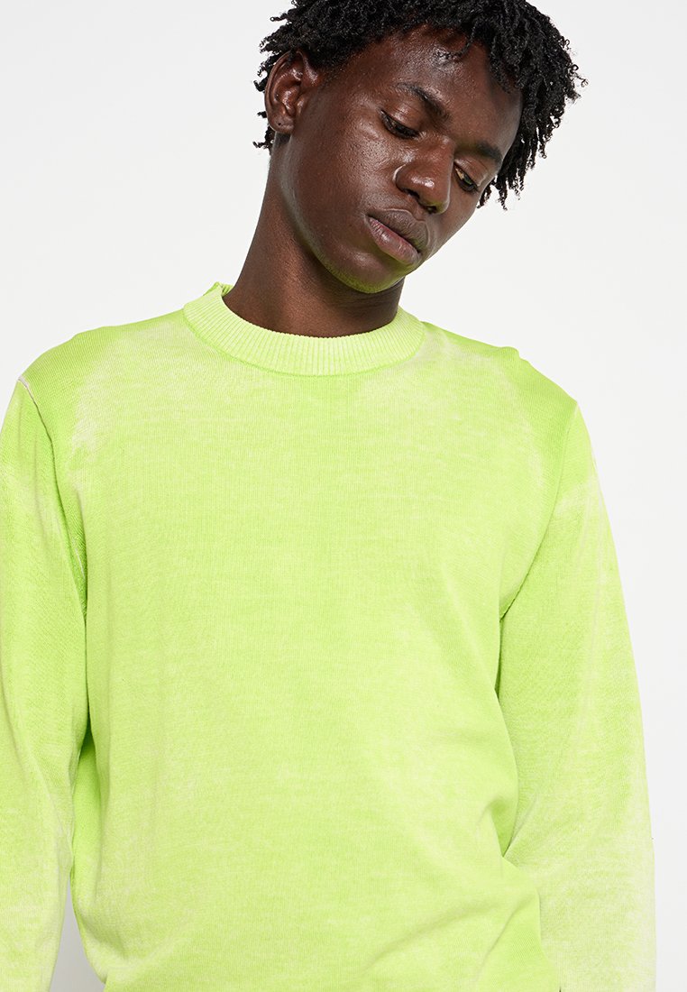 Junger Mann mit kurzen lockigen Haaren, der nach unten schaut, und einen leuchtend limegrünen Langarmpullover trägt, vor einem schlichten weißen Hintergrund.