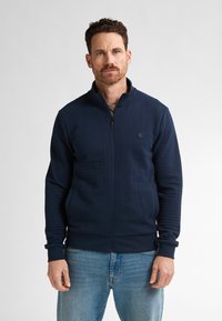 Námořnická bunda na zip s texturovaným materiálem, vysokým límcem a žebrovanými manžetami. Kombinována se světle modrými džíny. Jednoduchý design s decentním brandingem.