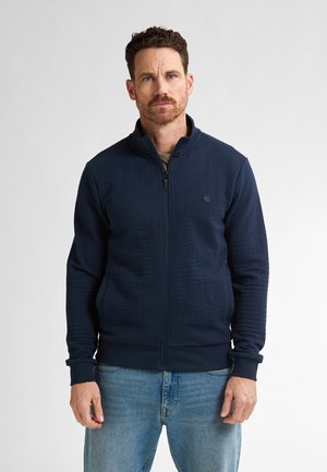 Marineblå zip-up jakke med tekstureret stof, høj krave og ribbede manchetter. Sammen med lyseblå jeans. Enkel design med subtil branding.