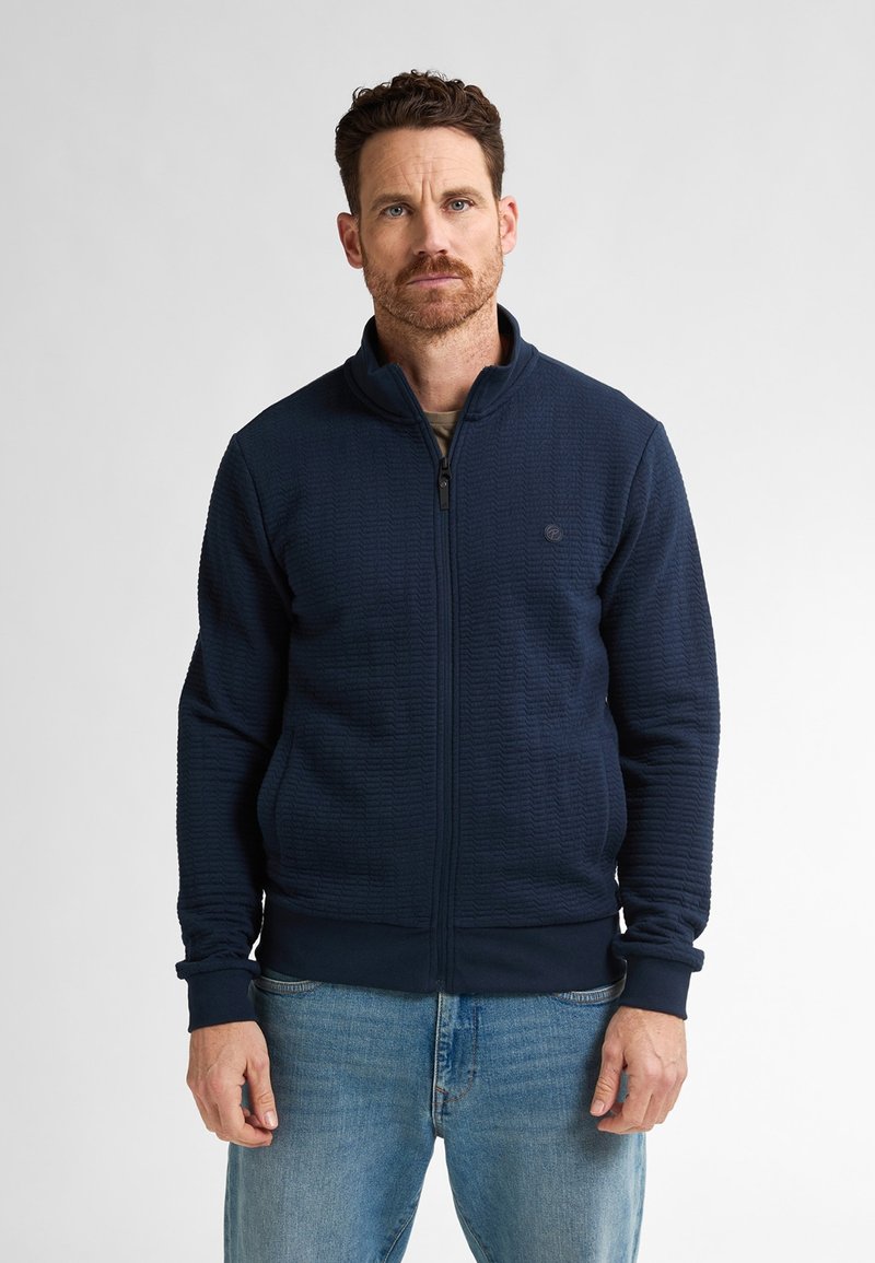 Námořnická bunda na zip s texturovaným materiálem, vysokým límcem a žebrovanými manžetami. Kombinována se světle modrými džíny. Jednoduchý design s decentním brandingem.