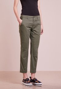 Pantalons cargo verts olive avec poches plaquées, jambes fuselées et fermeture à bouton. Portés avec des chaussures à enfiler à motifs.