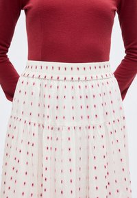 Γυναίκα που φοράει κόκκινο μπλουζάκι μακριάς μανίκι και άσπρη φούστα με ψηλή μέση και μικρό ροζ ανθικό σχέδιο, στέκεται με τα χέρια πίσω από την πλάτη.